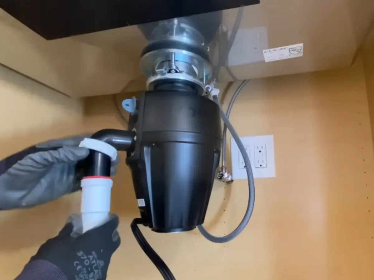 Gloved hands connecting a garbage disposal under a kitchen sink